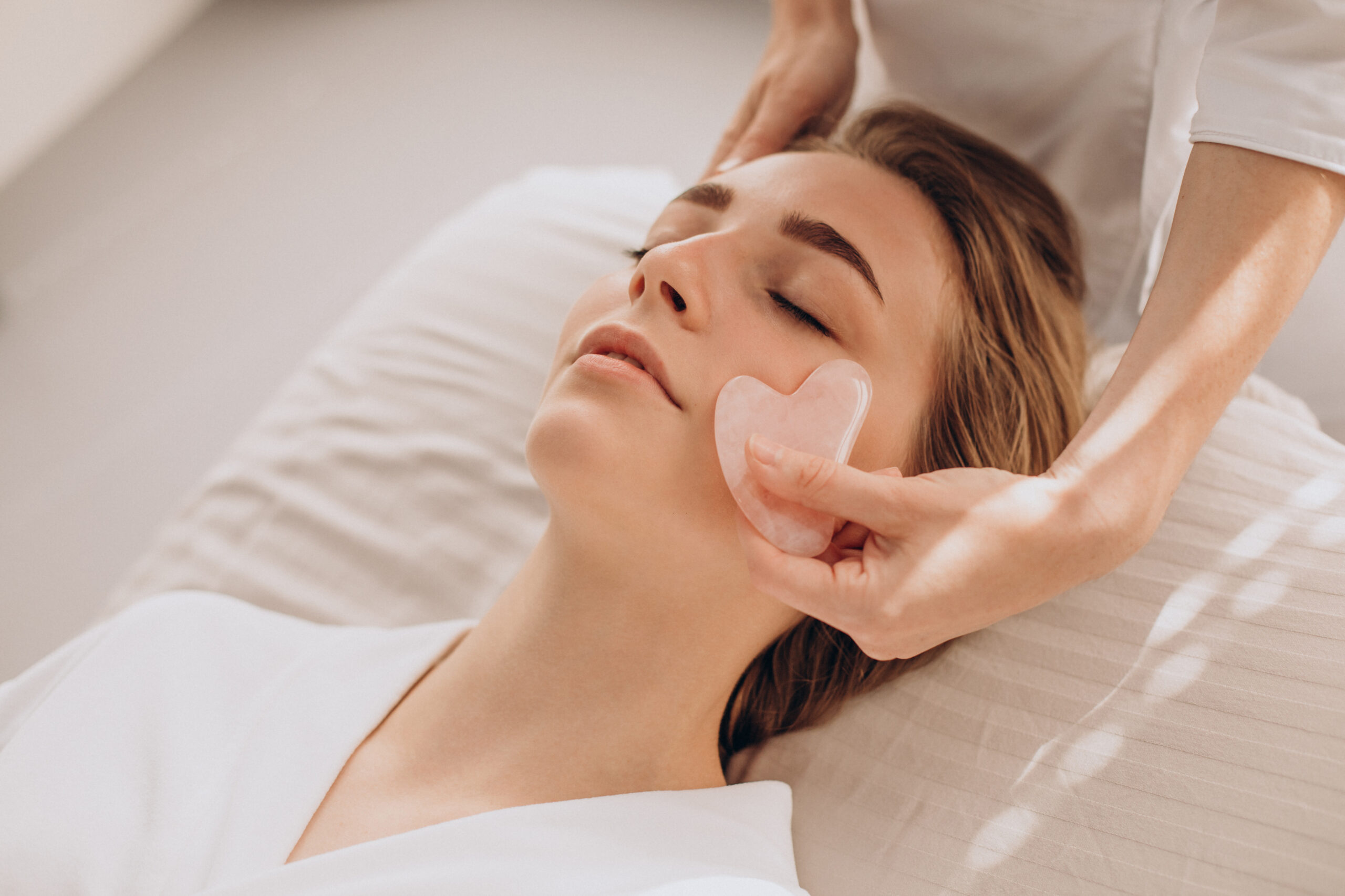 A spa treatment with a woman receiving a gentle facial massage using a rose quartz gua sha tool.